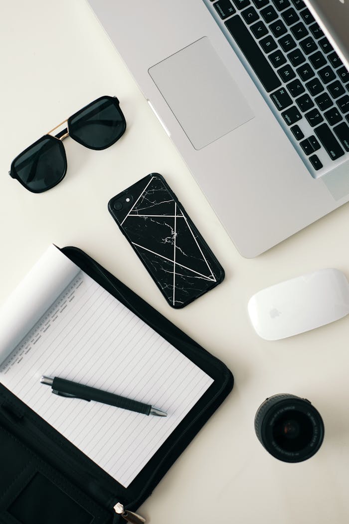 about-me A clean workspace featuring a laptop, smartphone, sunglasses, and stationery on a white surface.