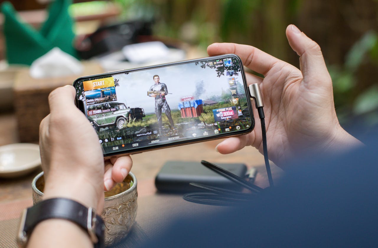 services-02 Person playing PUBG on smartphone outdoors with charging cable attached.